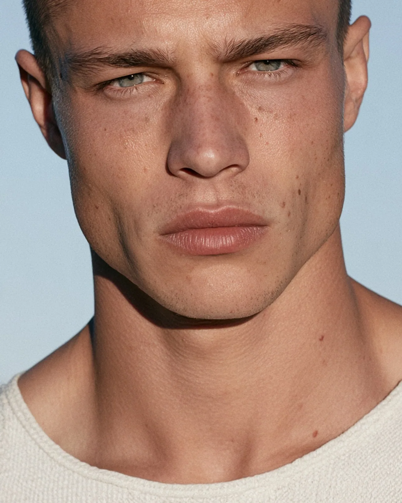 Close-up portrait of a young man with short dark hair, light eyes and freckles, wearing a cream knit top against a pale blue sky.