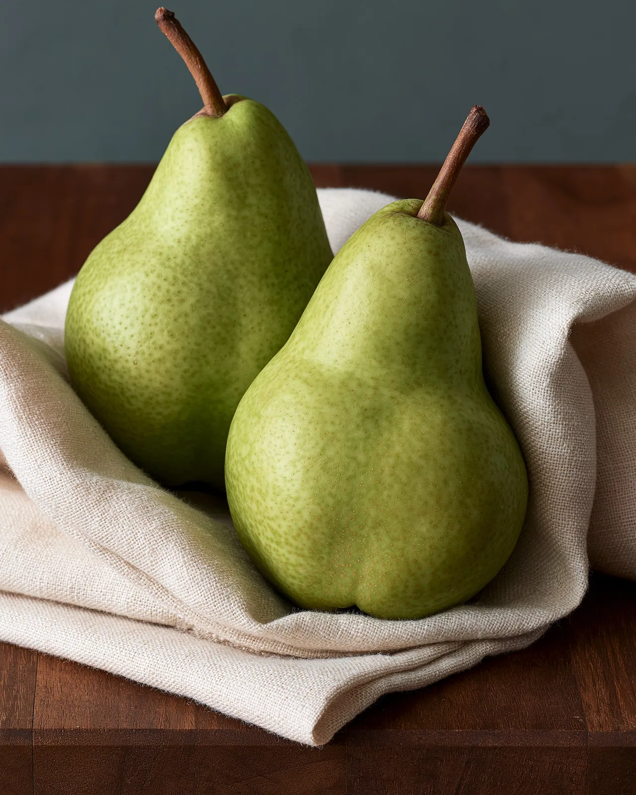 Photo of two green pears resting on a folded cream linen cloth on a dark wooden table against a slate gray wall.