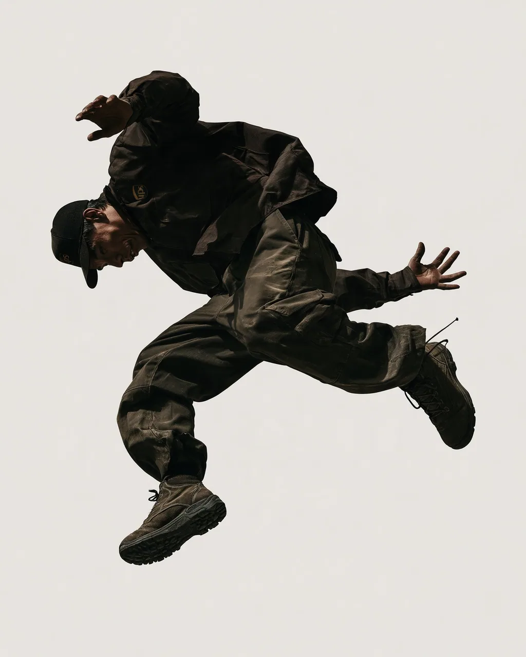 A man in a dark cap, jacket, cargo trousers and boots leaps sideways in midair against a plain off-white background.