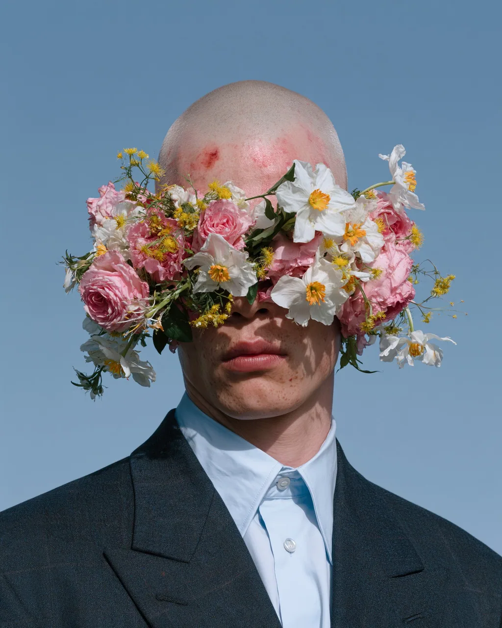 Color photo of a shaved-head person in a dark suit and pale blue shirt, pink and white flowers covering the eyes, against a blue sky.